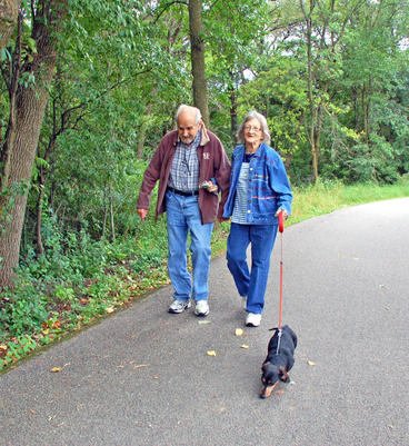 An older couple walking a dog on a paved walking trail